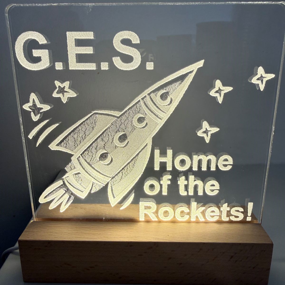Illuminated G.E.S. sign with a stylized rocket soaring among stars and “Home of the Rockets!” text; cheerful display on a wooden base.