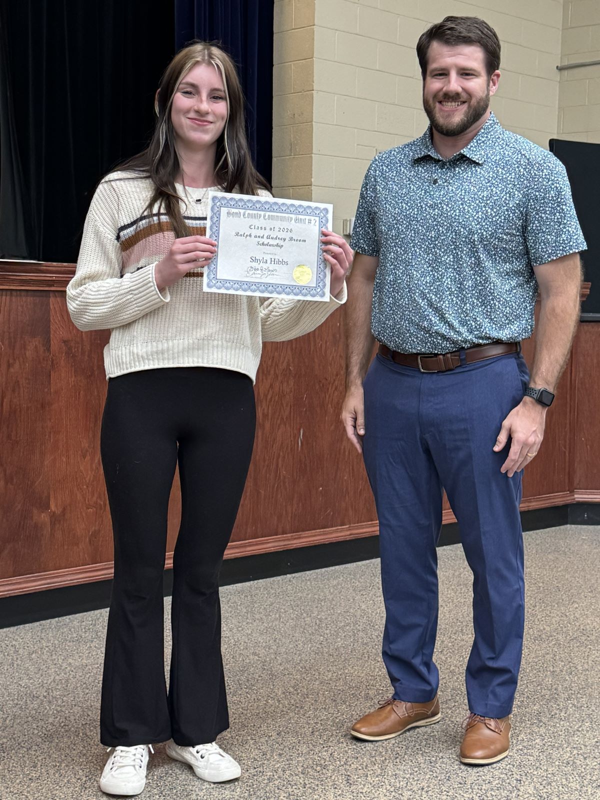 Broom Scholarship 2026 - Smiling female athlete and a male representing the AF pose indoors, holding a certificate, celebrating an award with a warm, proud atmosphere.