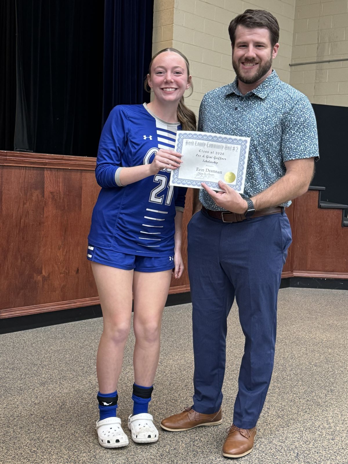 Gaffner Scholarship 2026 - Smiling student-athlete in blue jersey presents a certificate with a bearded man representing the AF in an auditorium, conveying pride and celebration.