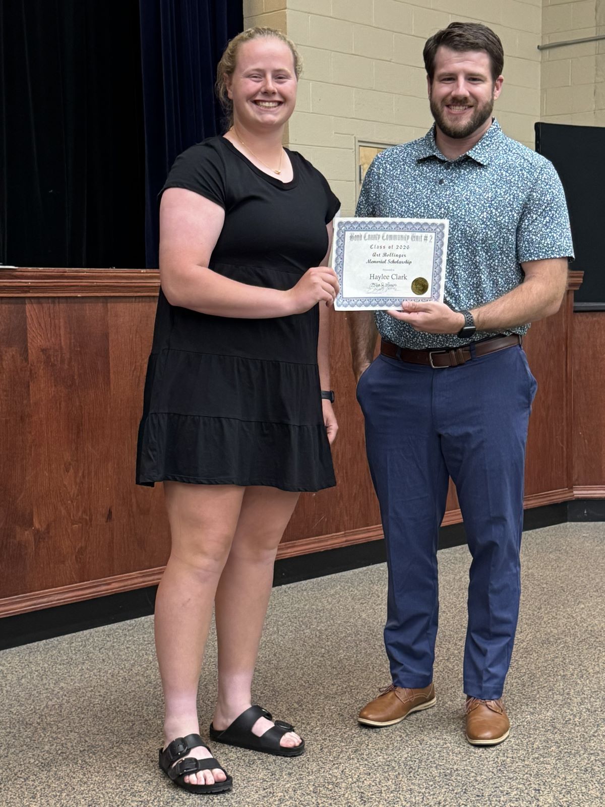 Bollinger Scholarship 2026 - Smiling young woman and a man representing the AF pose indoors, holding a certificate or award together during a recognition ceremony; warm, celebratory mood.