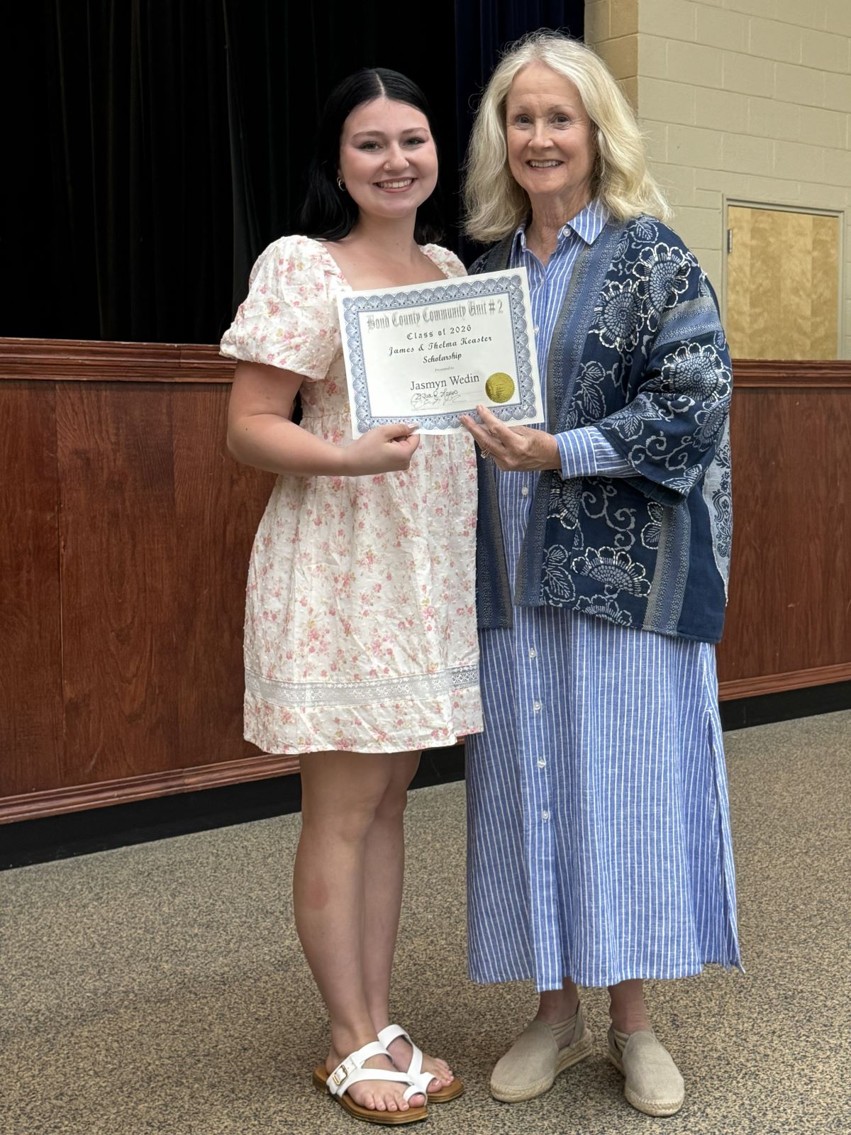 Keaster Scholarship 2026 - Smiling young woman and older woman representing the AF hold a framed award certificate together in an auditorium, conveying pride and celebration.