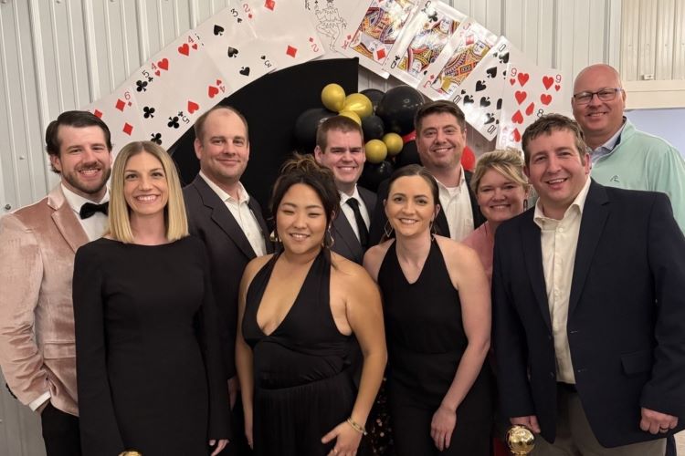 A smiling group of ten adults poses on a red carpet beneath playing-card arches and gold balloons at a festive casino-themed event.