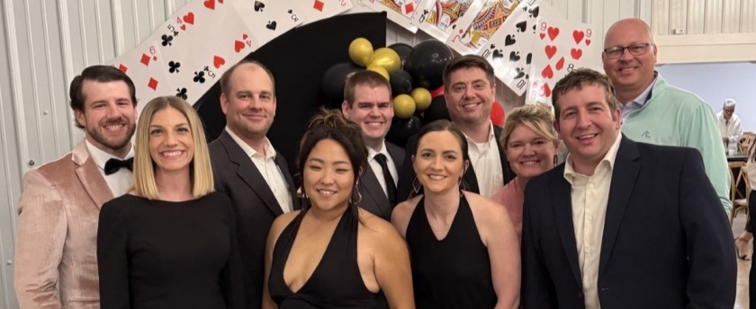 A smiling group of ten adults poses on a red carpet beneath playing-card arches and gold balloons at a festive casino-themed event.